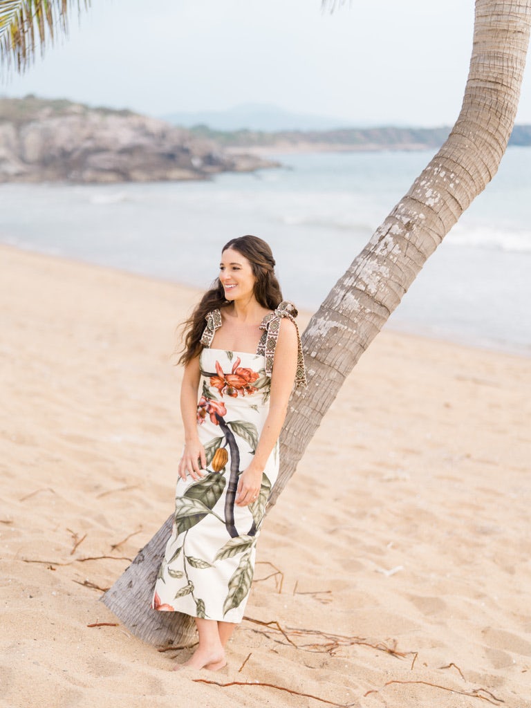 woman in a long white dress leaning against a palm tree on a beach