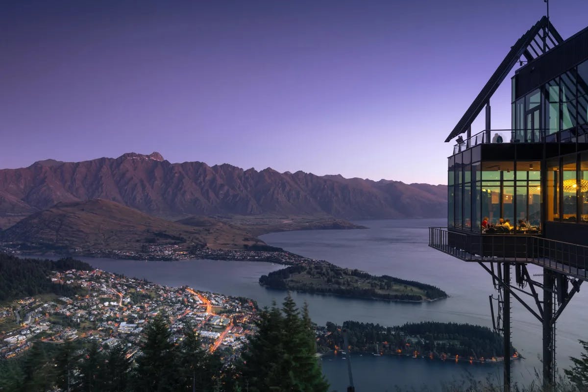 A beautiful image of Stratosfare Restaurant overlooking Lake Wakatipu, Queenstown. The sky is purple and the lodge is built on top of large stilts looking over the city, mountains and water. 