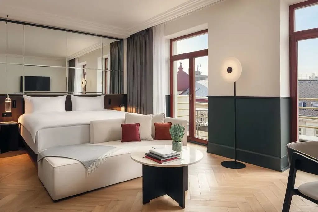 a sleek hotel room with a bed covered in white linens, a round coffee table, and wooden floors 