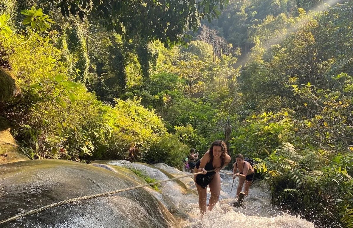 Two people hiking up a waterfall while pulling on a rope. There is a vast jungle in the background. 