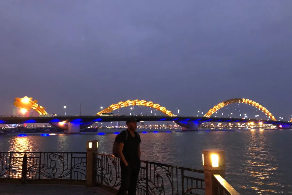 A picture of a man standing at the waterfront. At his back is the fully lit Dragon Bridge.
