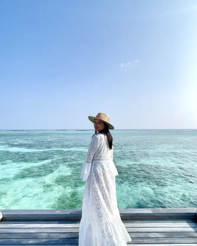 Cam wearing a white dress and hat in front of a view of crystal clear blue water
