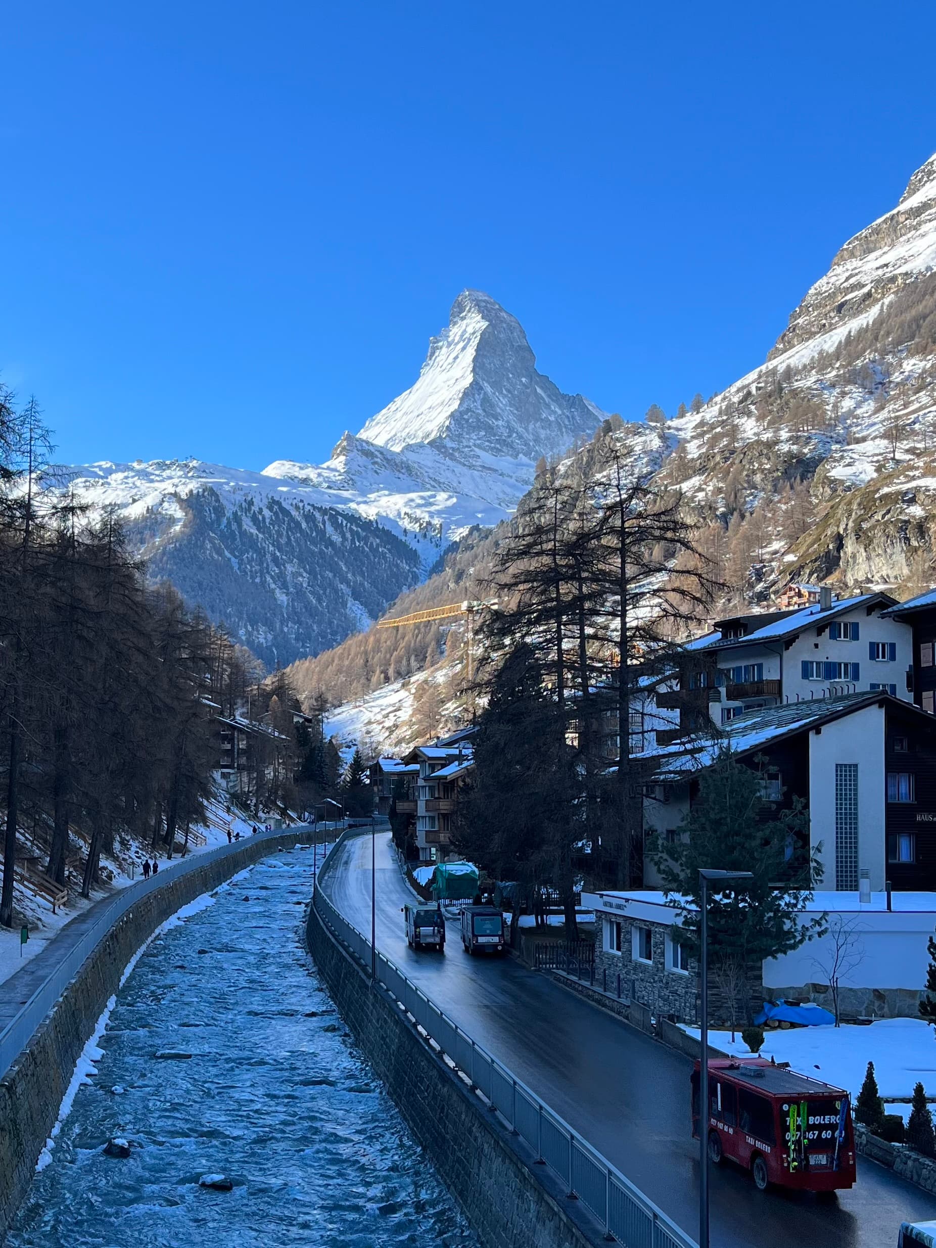 A snowy scene of Zermatt, Switzerland with a river, mountains, and quaint houses.