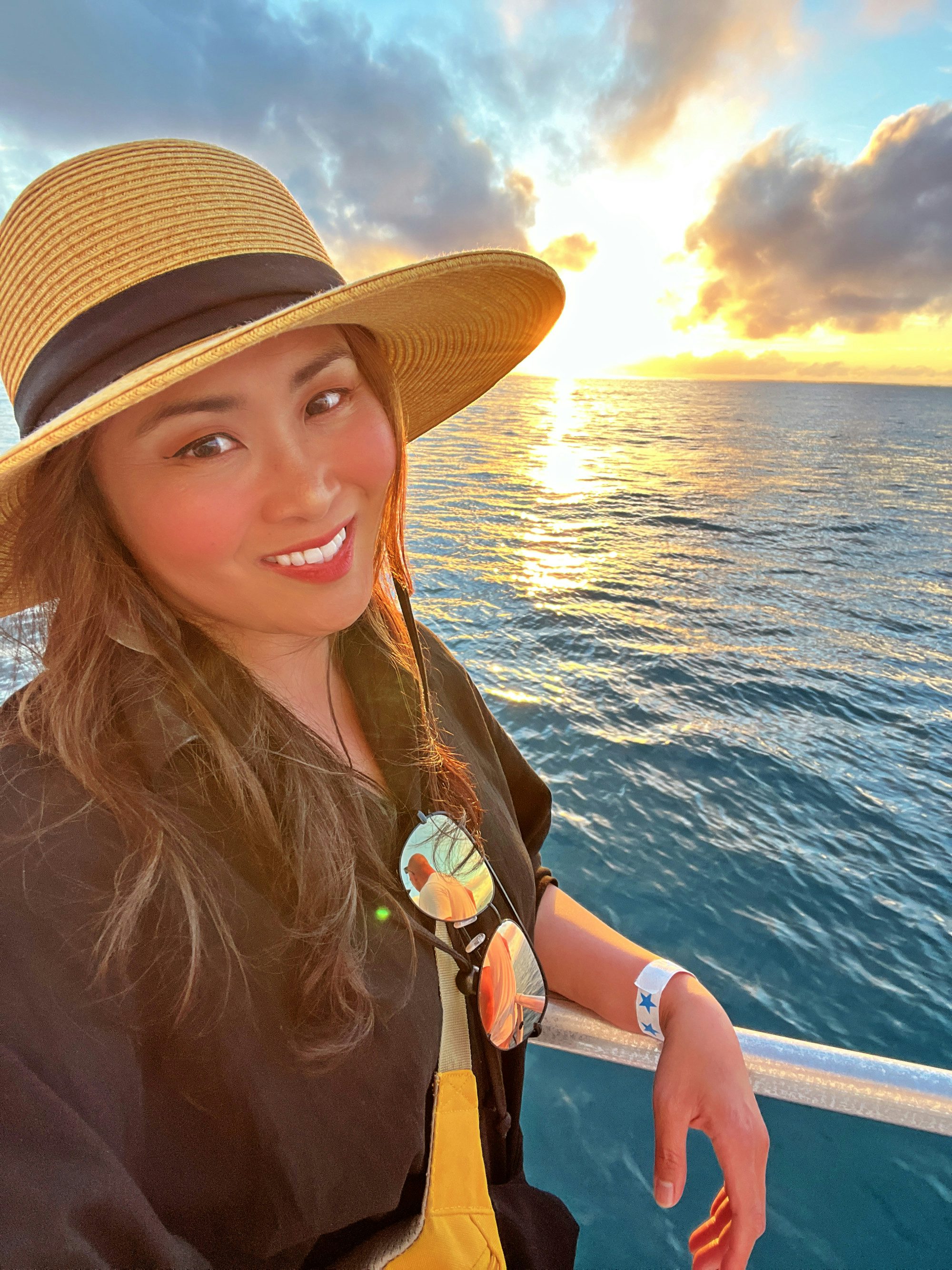 Travel Advisor Cheska Tariga leans on the railing of a boat cruising through the sunset wearing a sun hat