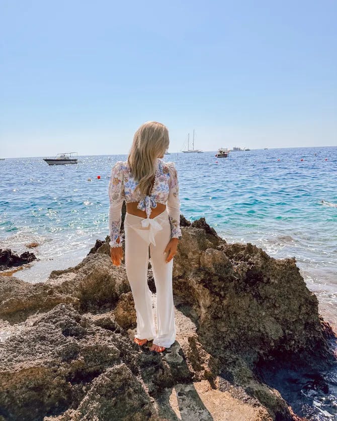 A picture of Morgan wearing white pants and a cropped top while standing on a rock looking back at the blue ocean 