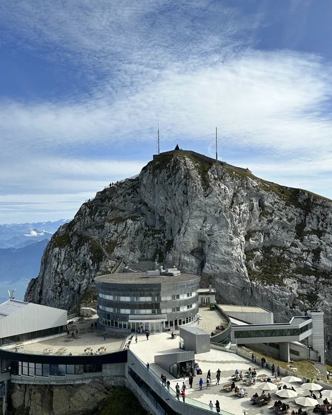 An aerial view of Mount Pilatus with people walking near a grey building built into the mountainside with blue sky and clouds in the background