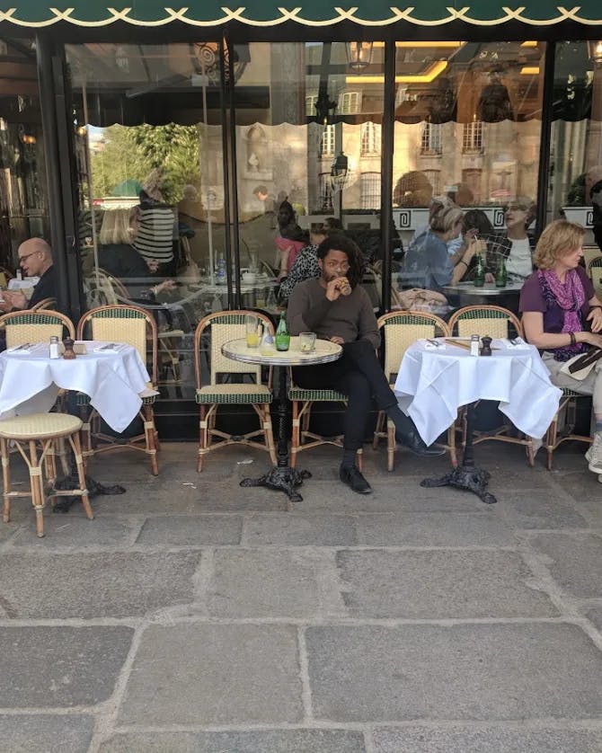 People dining on tables and chairs on a city street in front of windows and under a balcony 