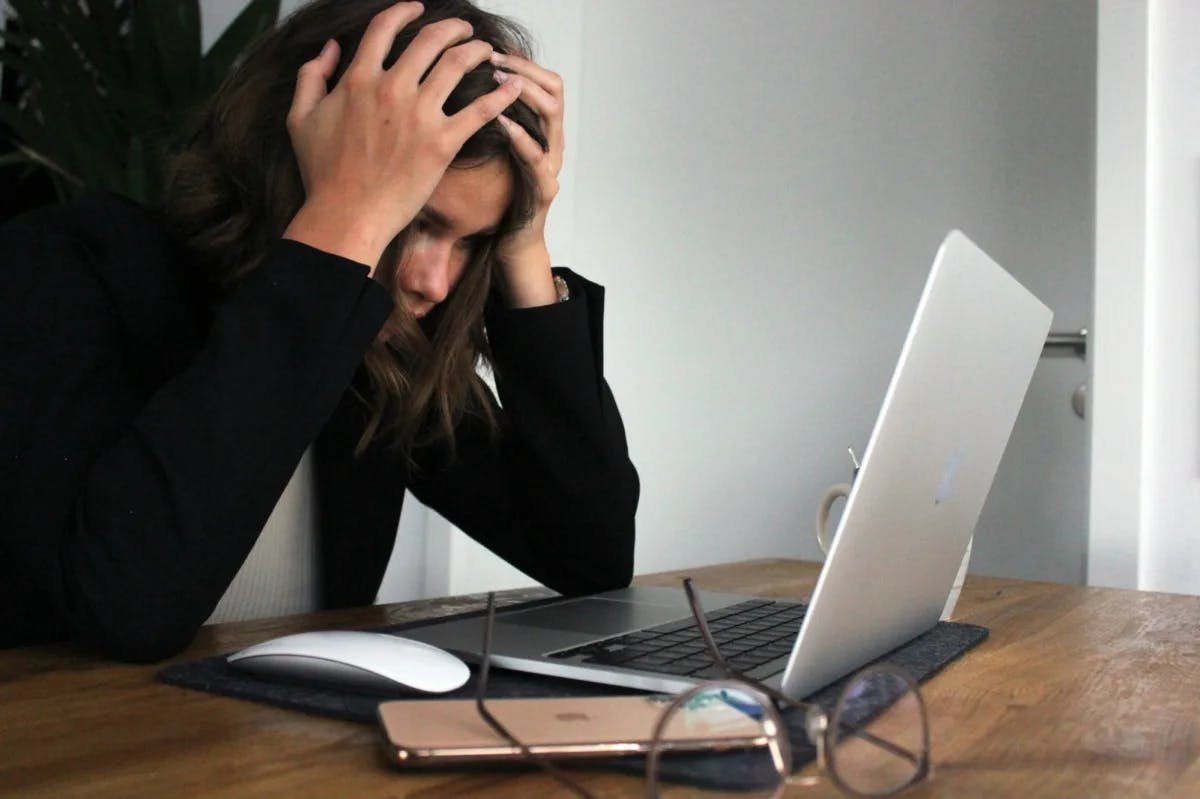 A young woman holds her head in frustration while staring at her laptop on a wooden desk