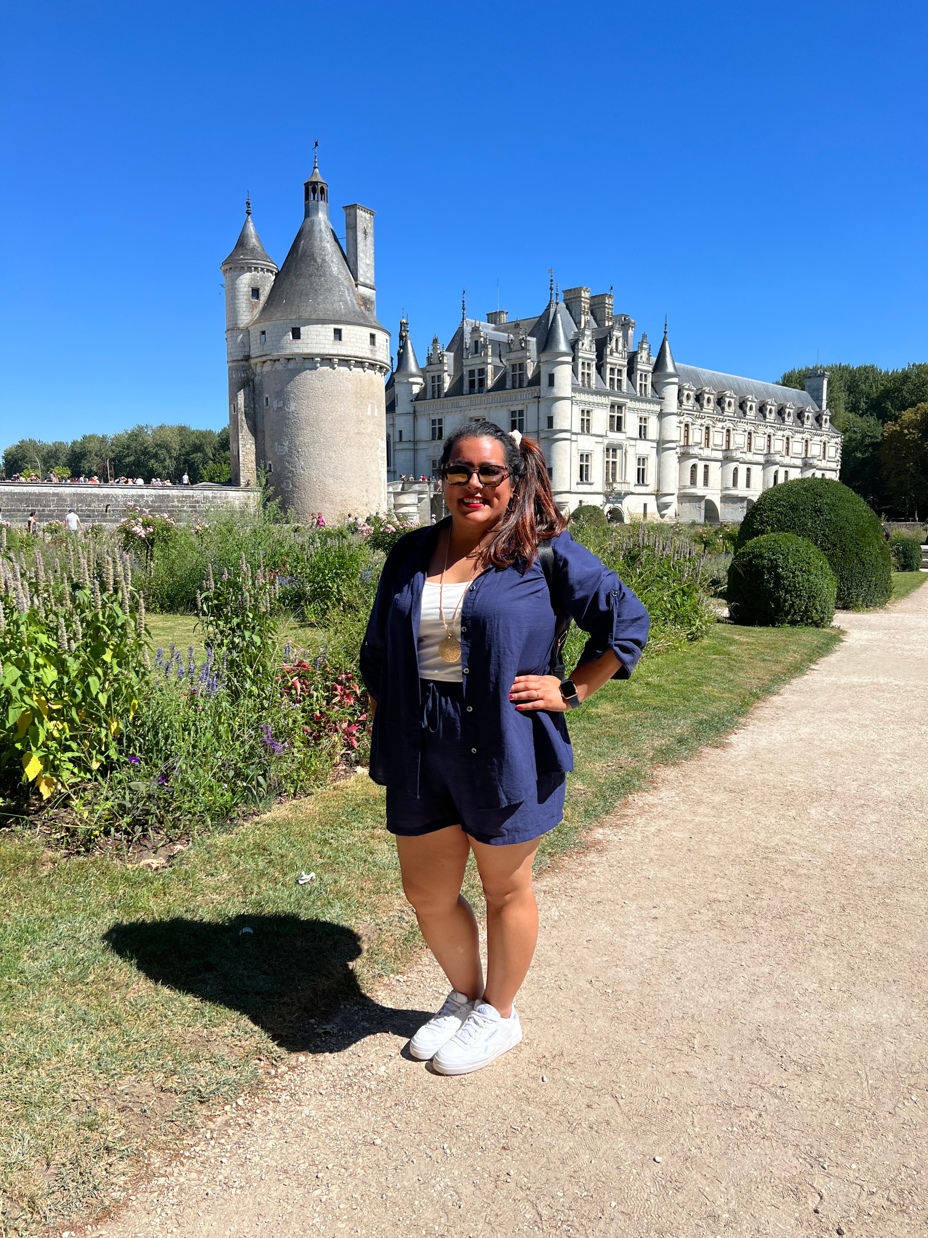 Pooja posing and smiling in front Château de Chenonceau castle on a sunny day in France.