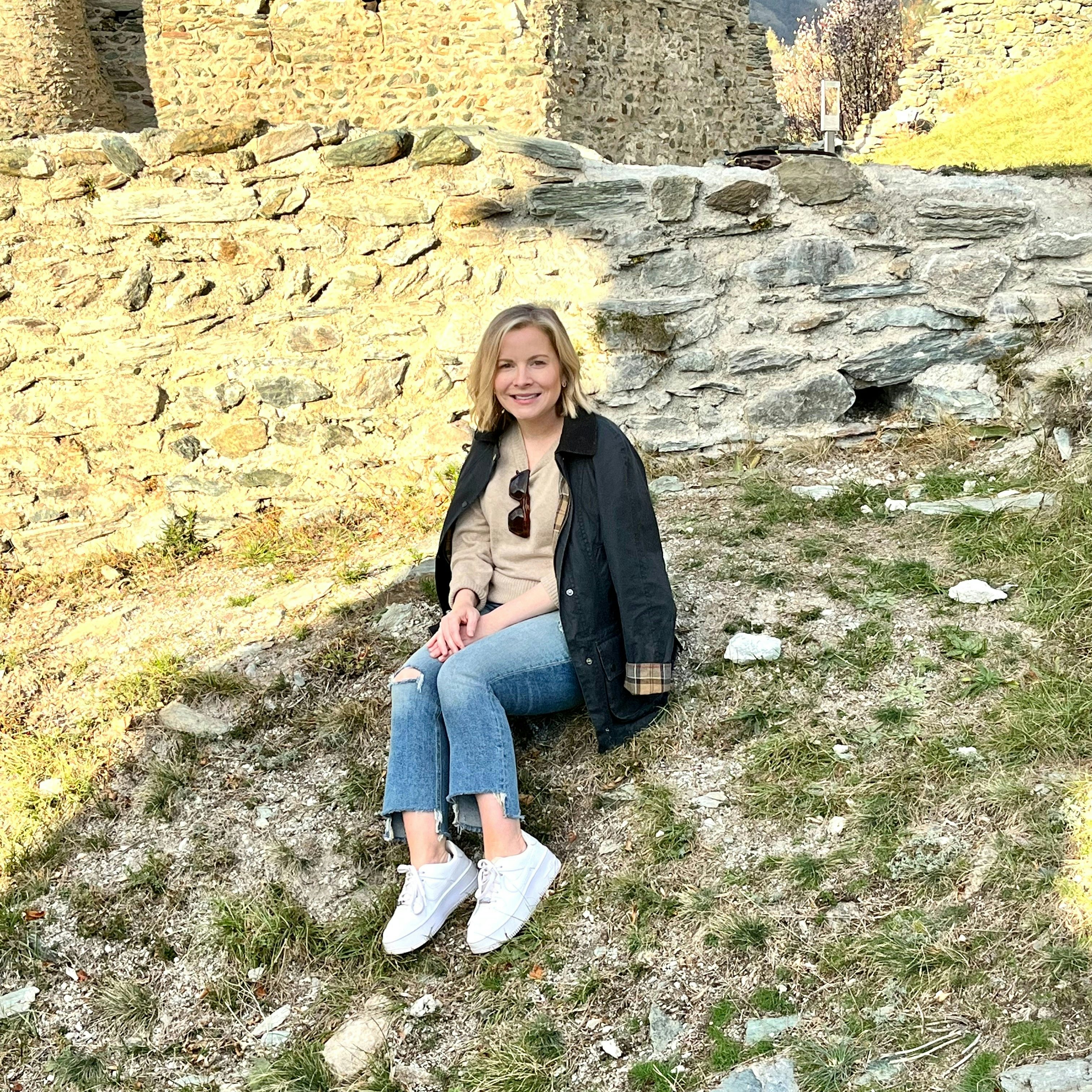 Travel Advisor Alyson Ward in jeans and a tan top with a black coat sitting on a green grass and stone covered hill.