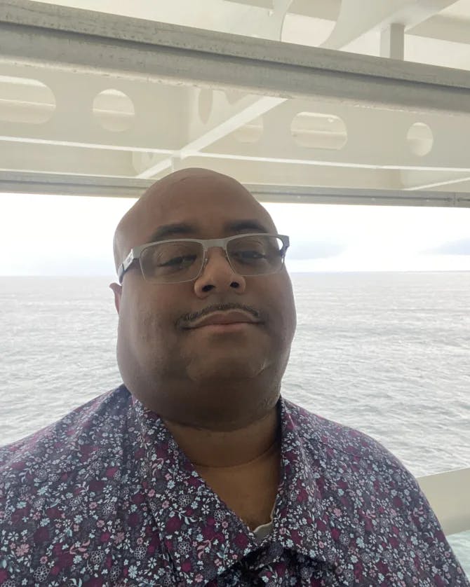 Mario posing for a selfie while wearing a patterned blue top with the grey ocean water in the background on a cloudy day.