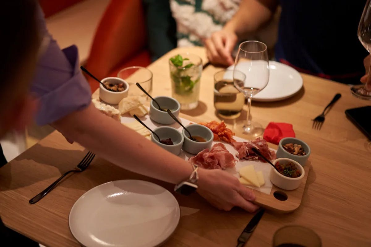 A wooden table full of plates of food, silverware and wine glasses. There is a person holding a wooden charcuterie board with various toppings placing it down onto the table. 
