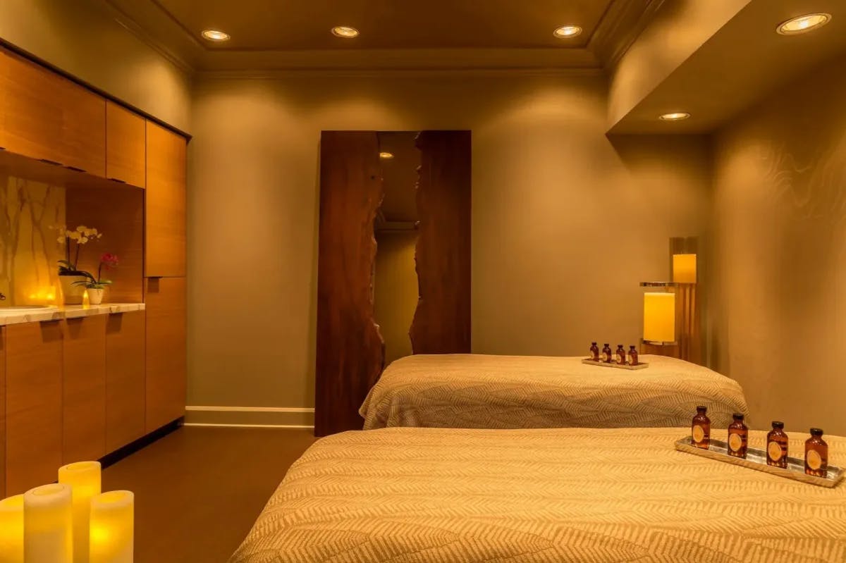 A tranquil spa room lit with warm lighting and various oils sitting on tray on twin massage tables