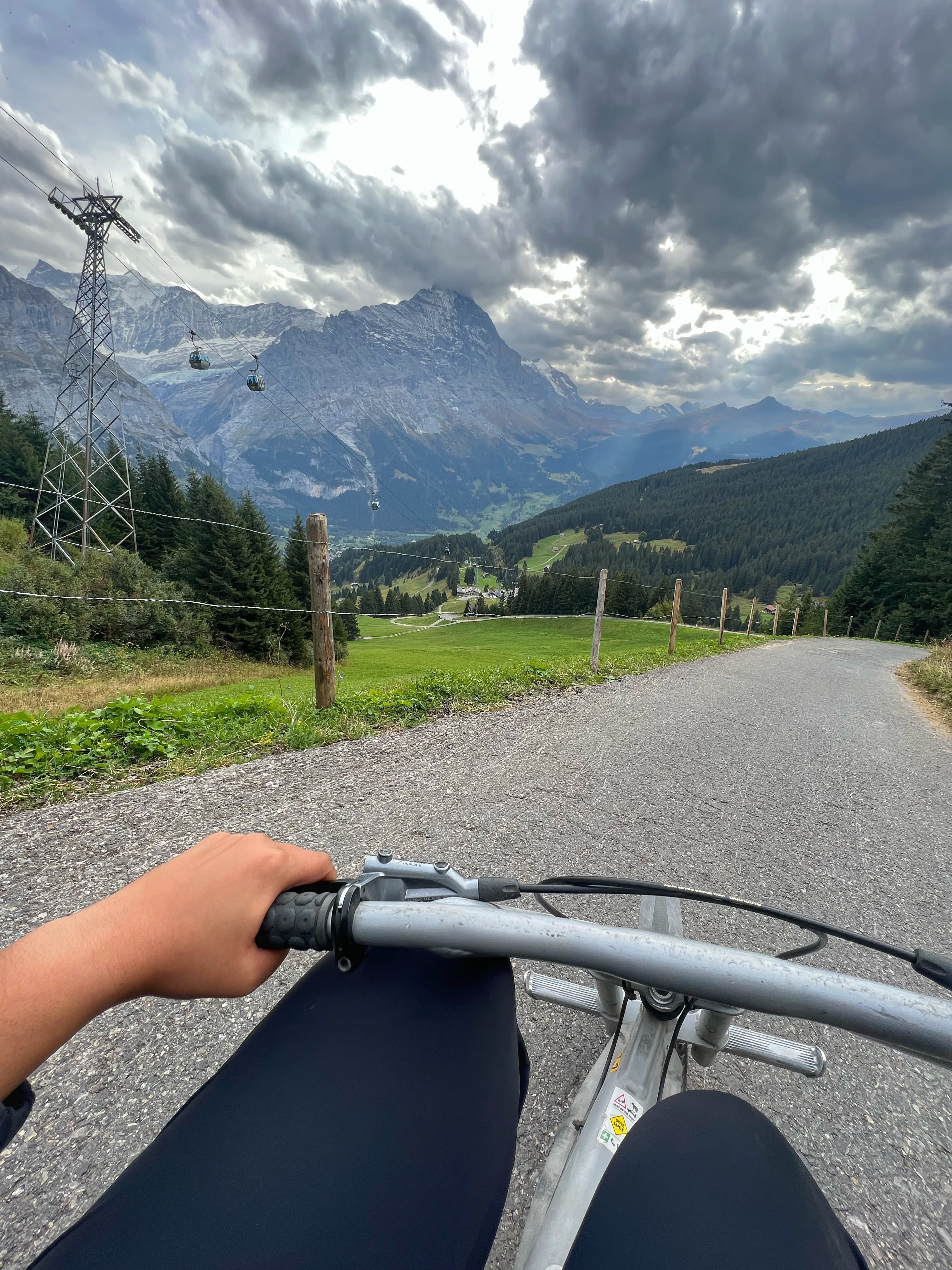 Point of view photo from the handlebars of a bicycle with a country road, mountains, and a cloudy sky in front.