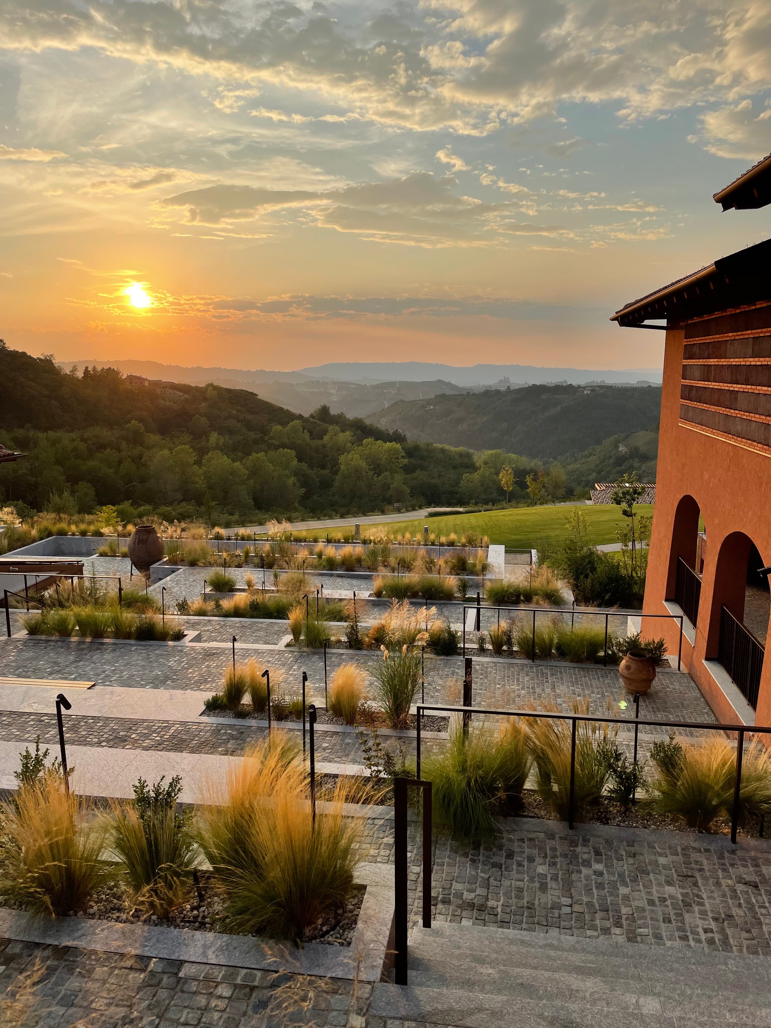 A sunset view of Casa di Langa surrounded by landscaping, rolling hills and a rusty colored building