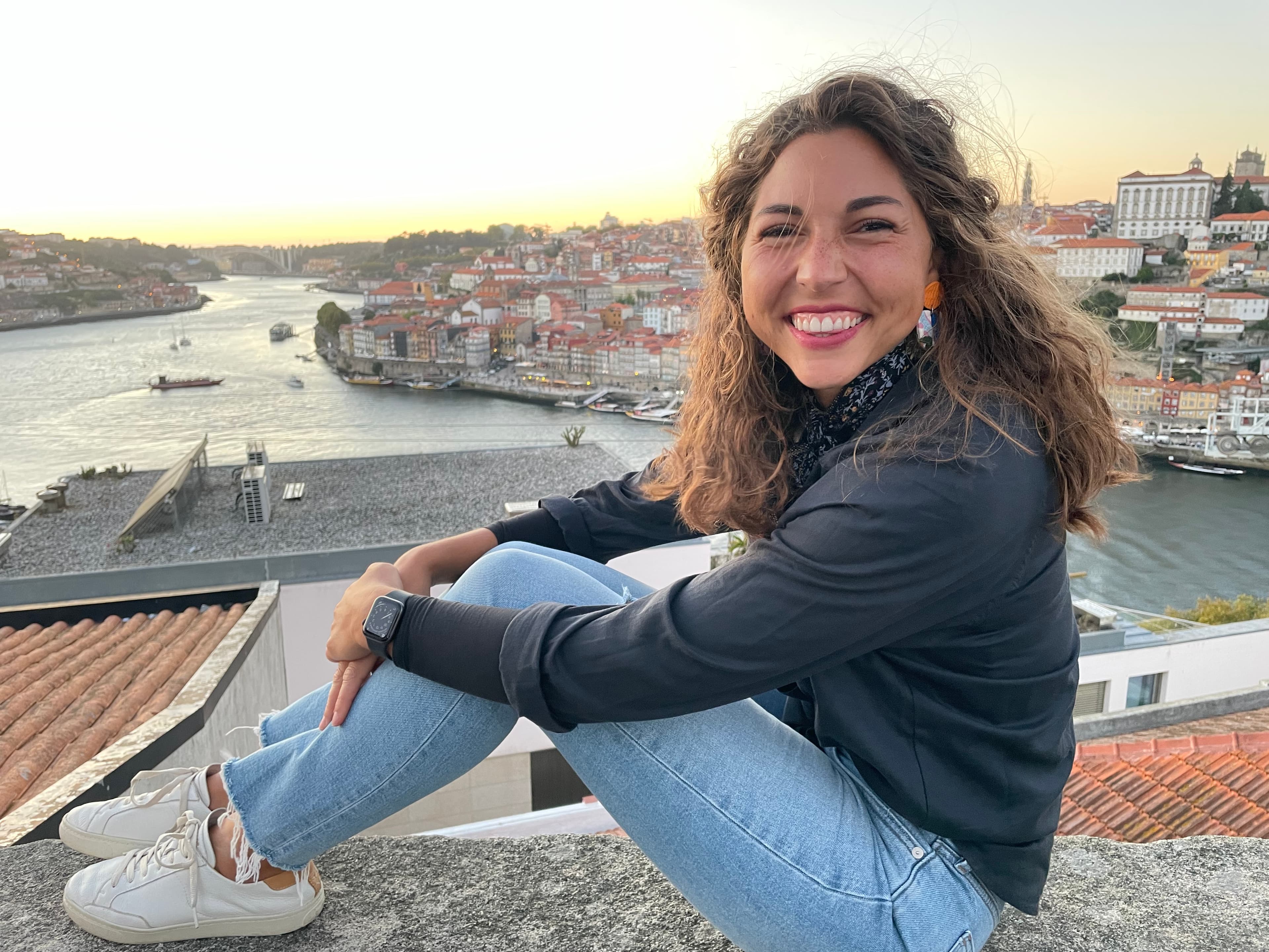 Emily wearing a grey top and blue jeans while posing in front of a river and buildings at sunset.