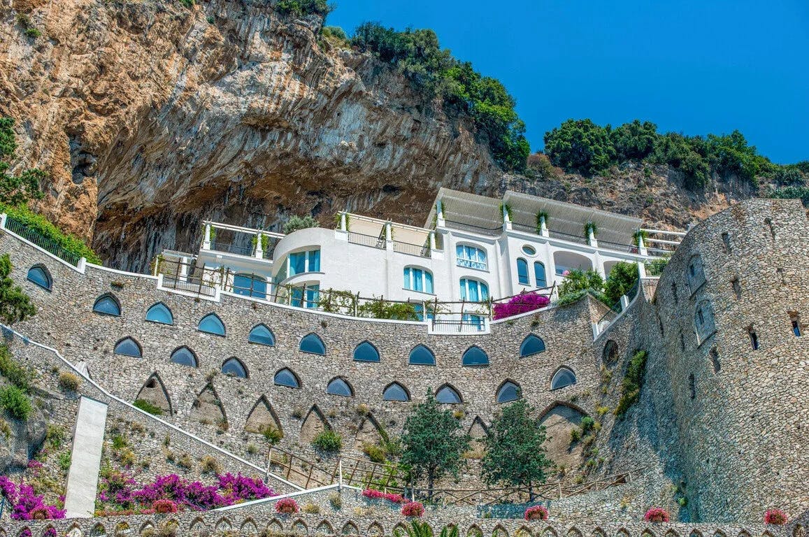 From the shore (unseen), the steep, tiered walls of Borgo Santandrea, with rooms built into the cliffside. At the top, a whitewashed hotel stands prominently, surrounded by manicured gardens