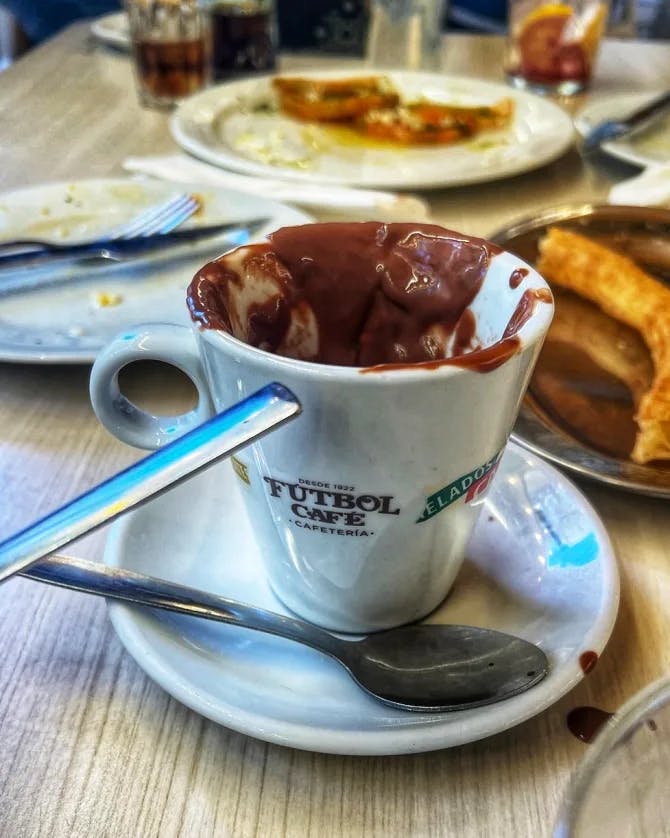 A white mug and saucer full of chocolate with a silver spoon resting on the side and pastries on three plates in the background