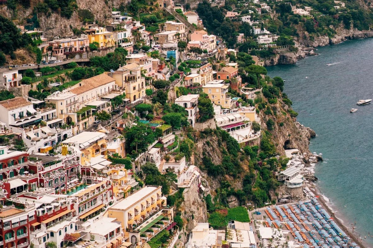 Rows of historic structures stand above a filled beach along the face of a cliff in Positano, Italy