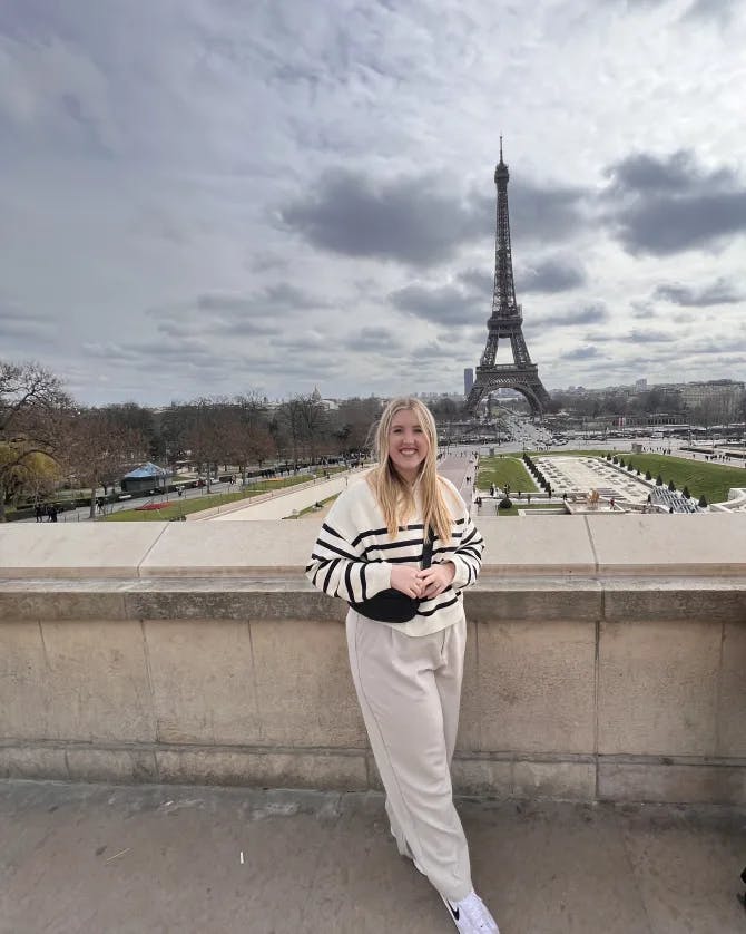 Alyssa wearing a black and white sweater and cream colored trousers posing on a sidewalk in front of the Eiffel Tower in Paris, France