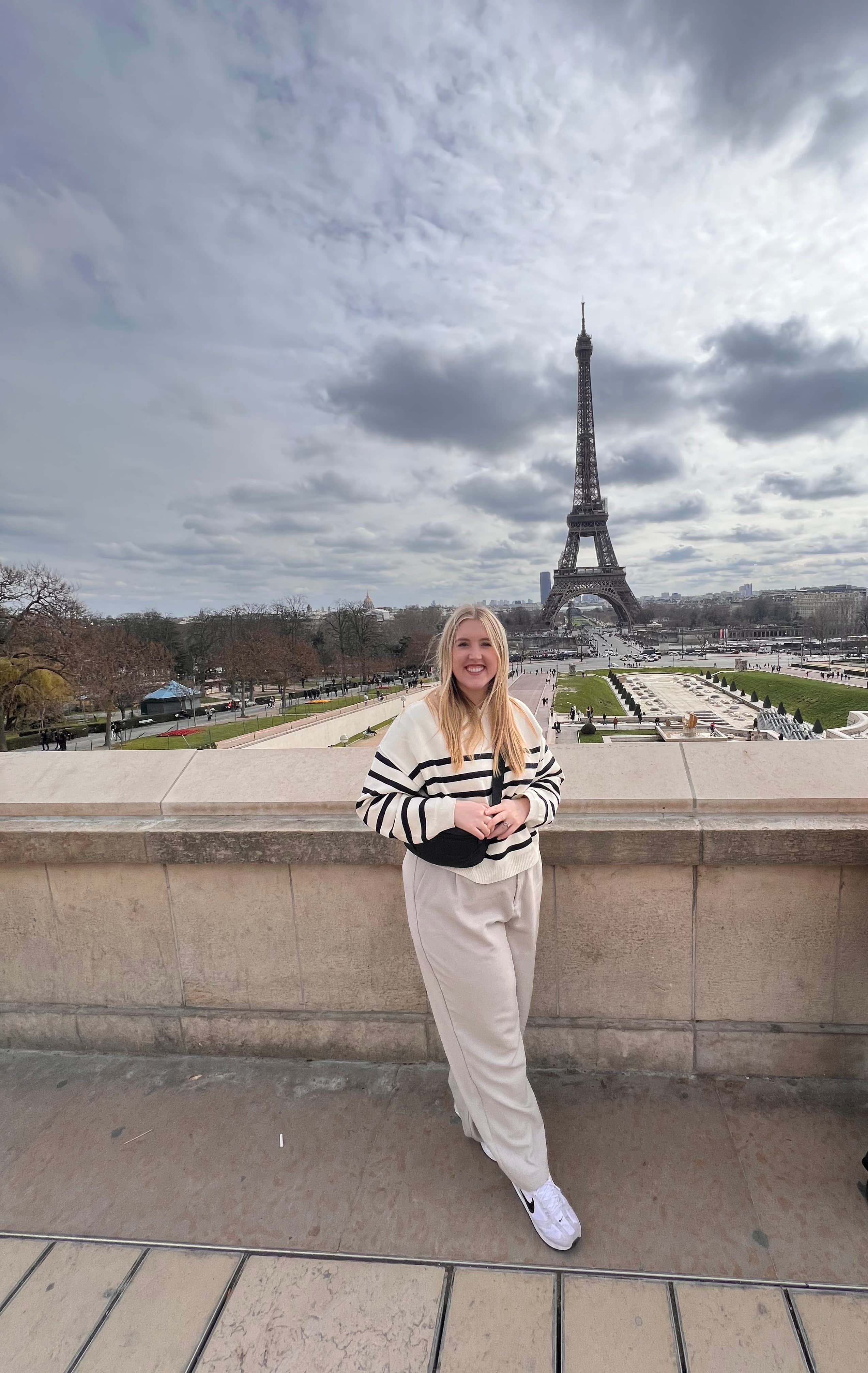 Alyssa wearing a black and white sweater and cream colored trousers posing on a sidewalk in front of the Eiffel Tower in Paris, France