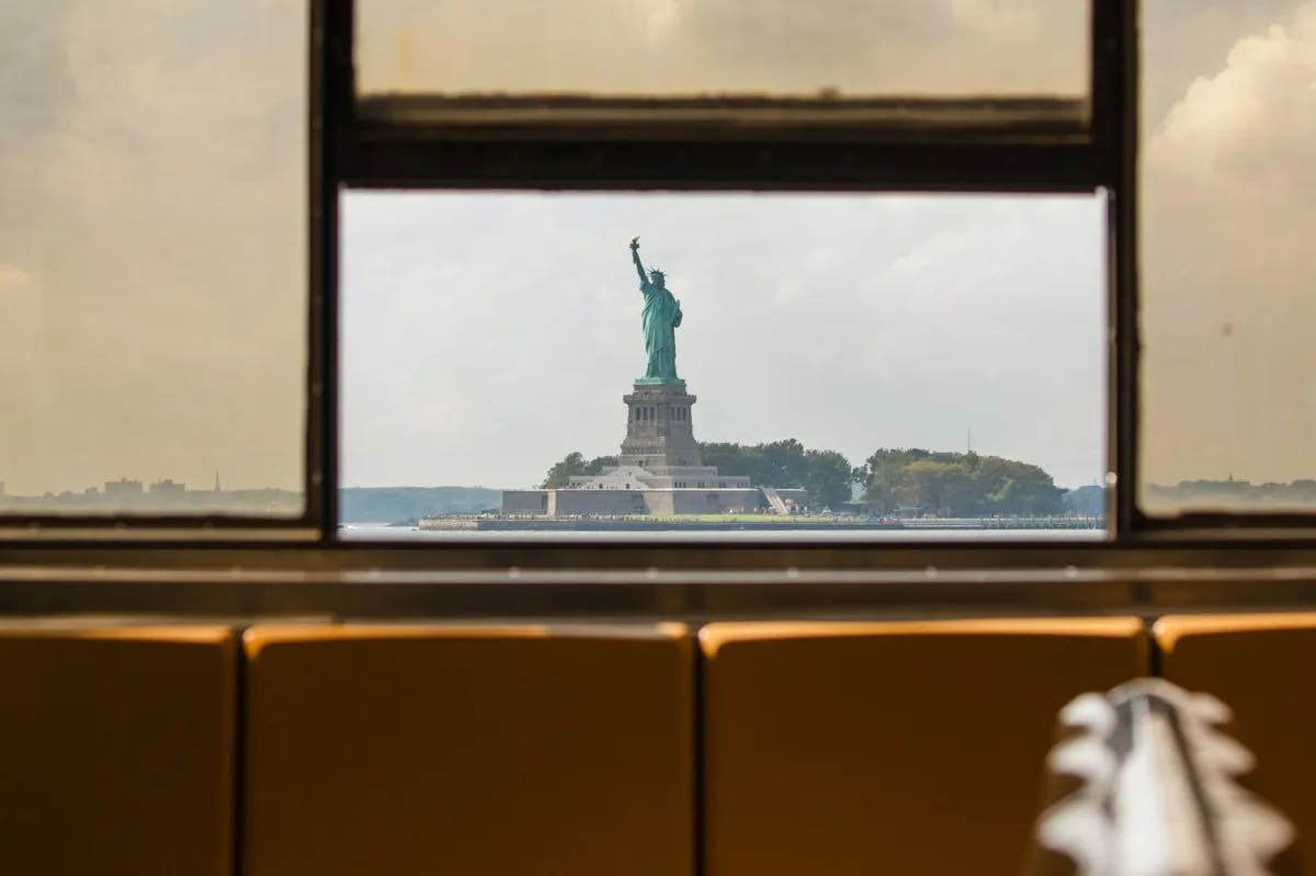 The Statue of Liberty stands prominently in the foreground from a view through a window on the Staten Island Ferry