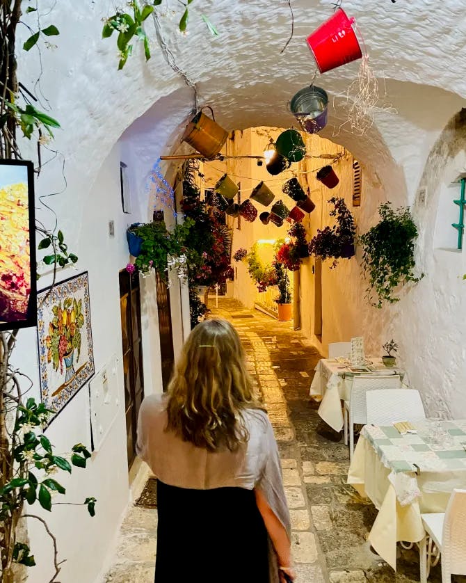 Jennifer walking down a stone pathway under a white arched ceiling decorated with pots and plants while dining table align the side of the wall