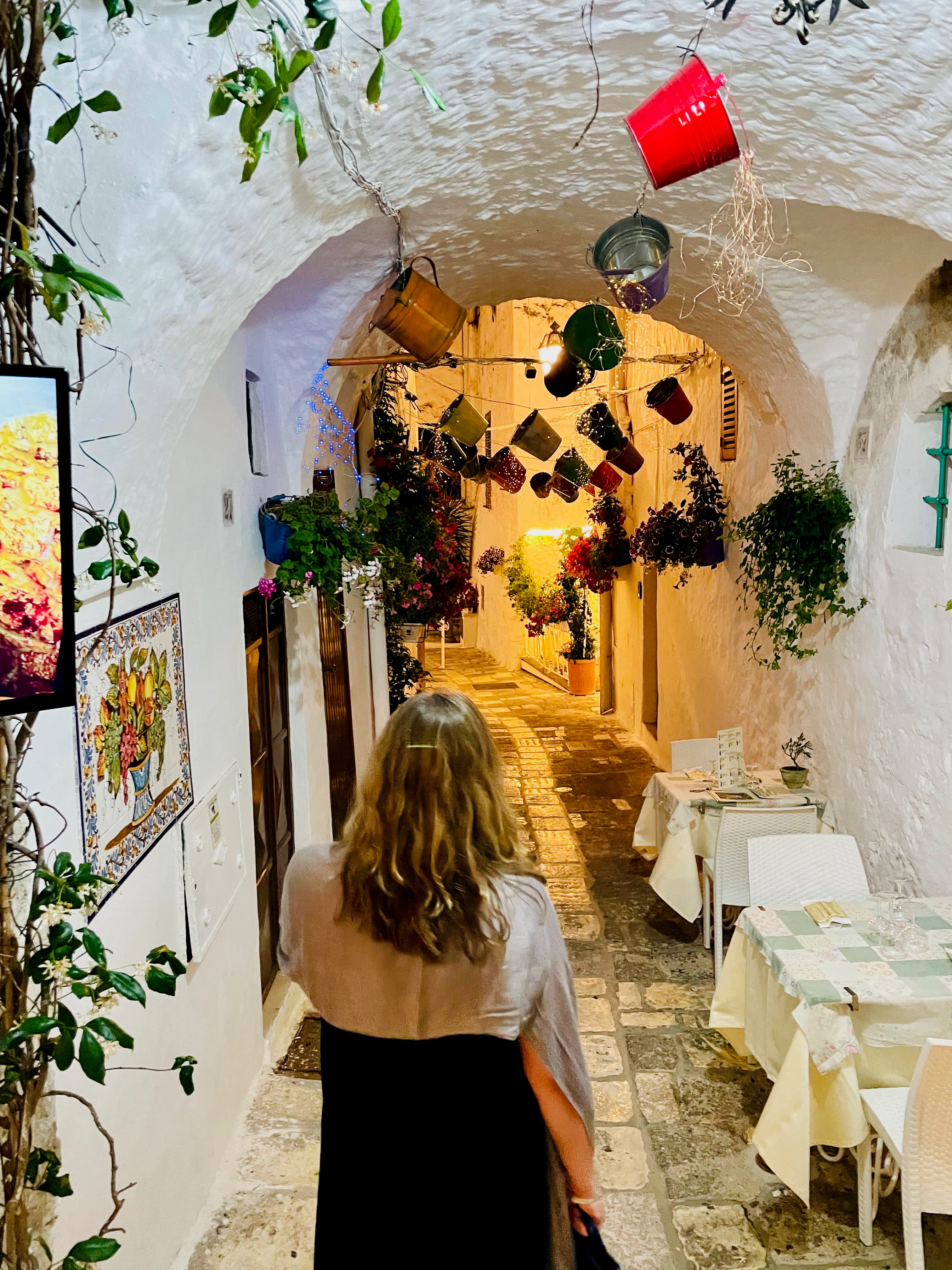 Jennifer walking down a stone pathway under a white arched ceiling decorated with pots and plants while dining table align the side of the wall