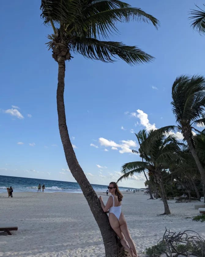 A picture of Sonia wearing a swimsuit and leaning against a palm tree on the beach with the ocean in the background