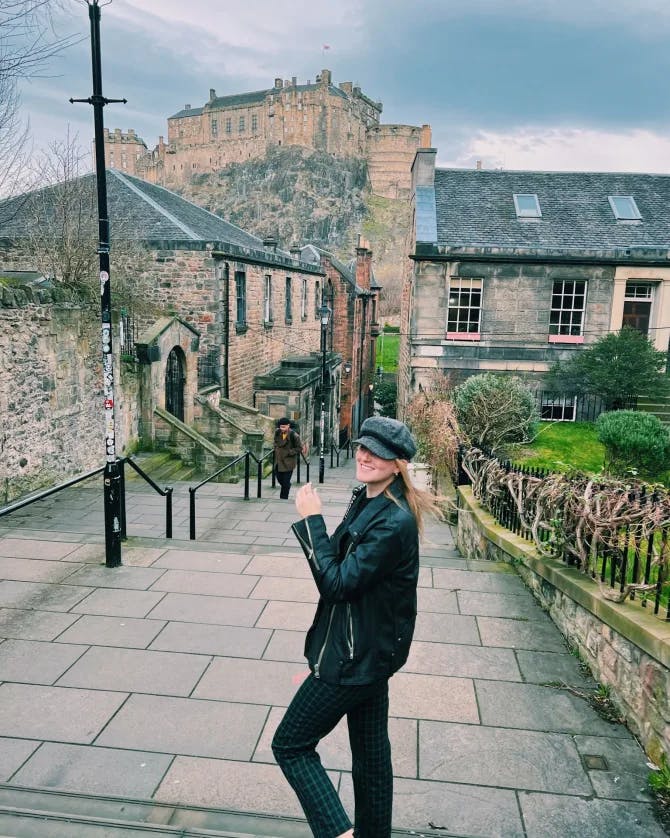 Picture of Courtney in black jacket in Edinburgh with a cute village and stone pathways behind her.
