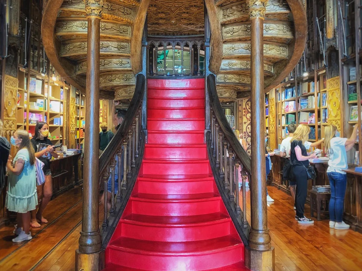 The image depicts an ornate, wooden staircase with a vibrant red carpet in the center of a beautifully decorated bookstore. 