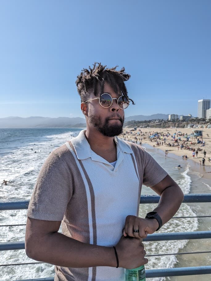 Norris posing in a white and tan polo top on a boardwalk in front of a busy beach and ocean