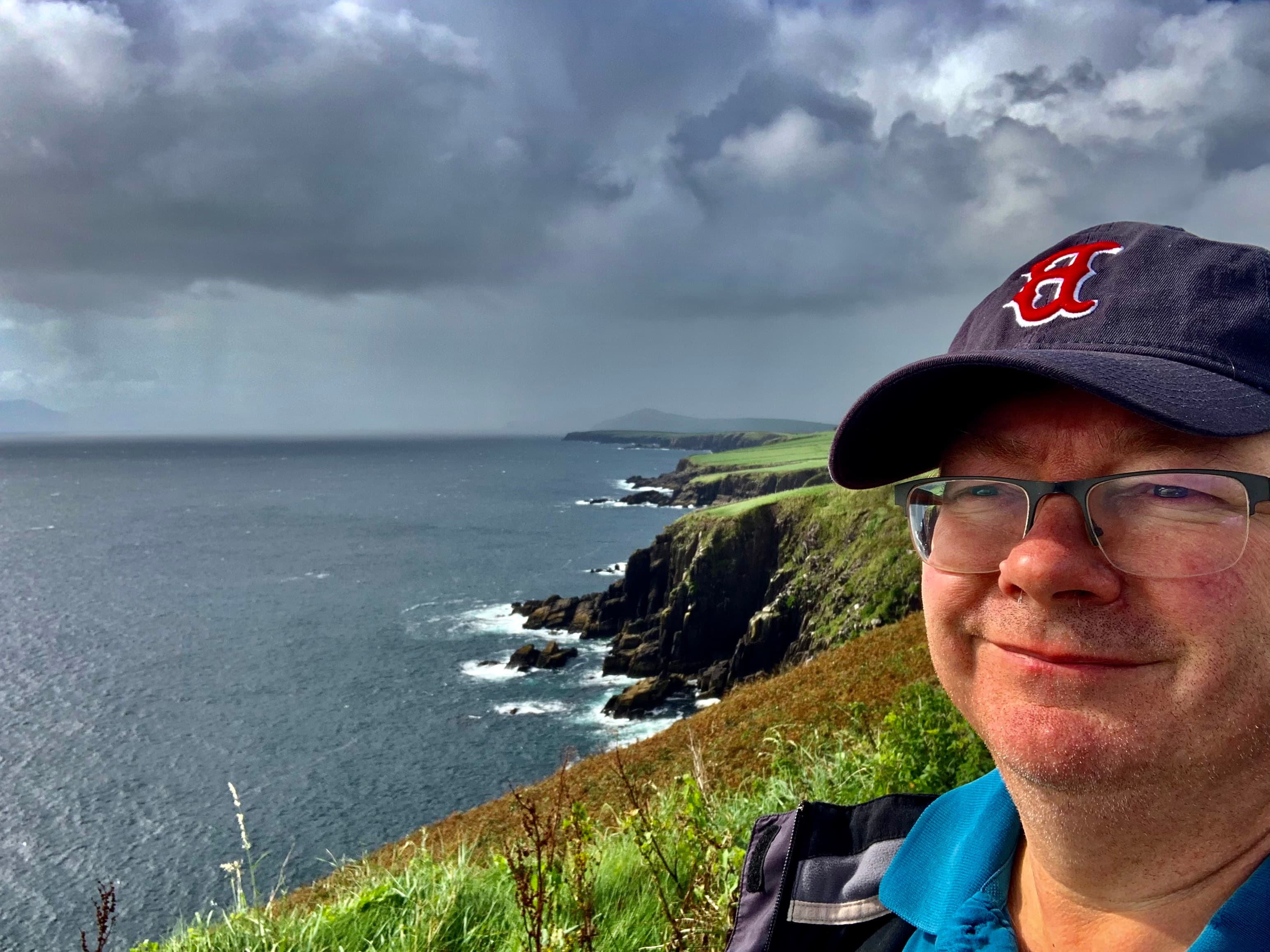 Pat standing by the edge of a grassy cliff, which descends into the sea, on a cloudy day.
