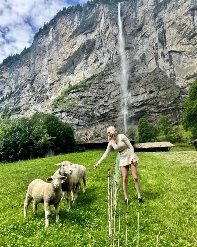 Picture of Alycia standing on green grass and leaning in to pet sheep in front of Staubbach Waterfall 