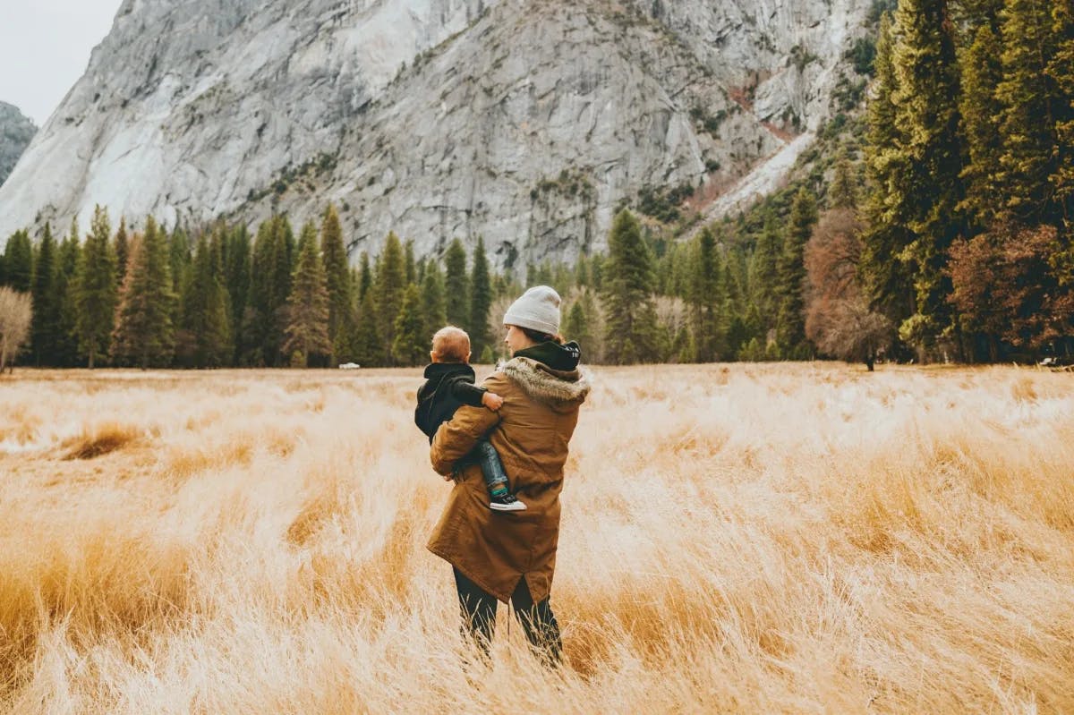 A woman in warm attire walks through scenic plains with mountains in the distance while holding her young boy