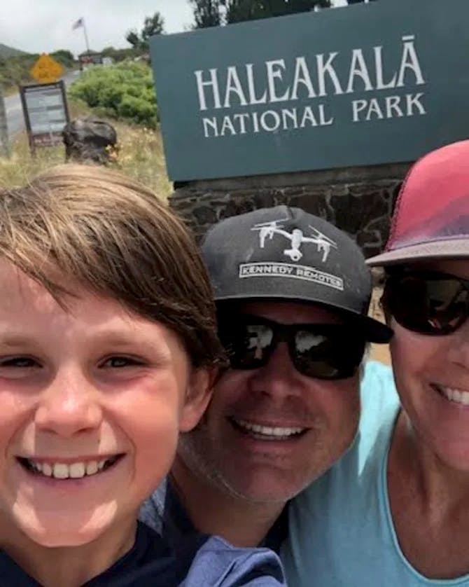 Three people posing for a selfie in front of the Haleakala National Park sign with a white parked car and road to the left