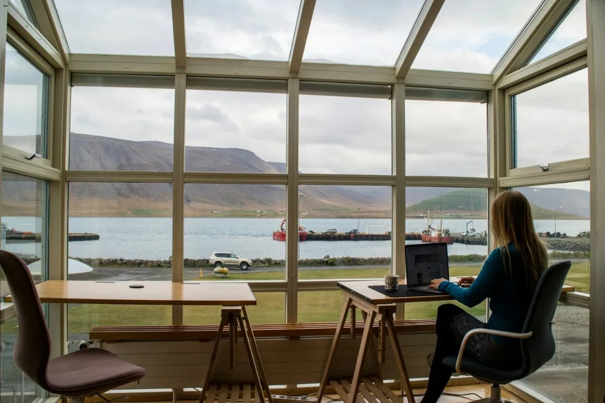A lone woman works at a coffee shop table with an immediate view of an Icelandic landscape: dreary green fields lead to a body of water separating more fields and mountains