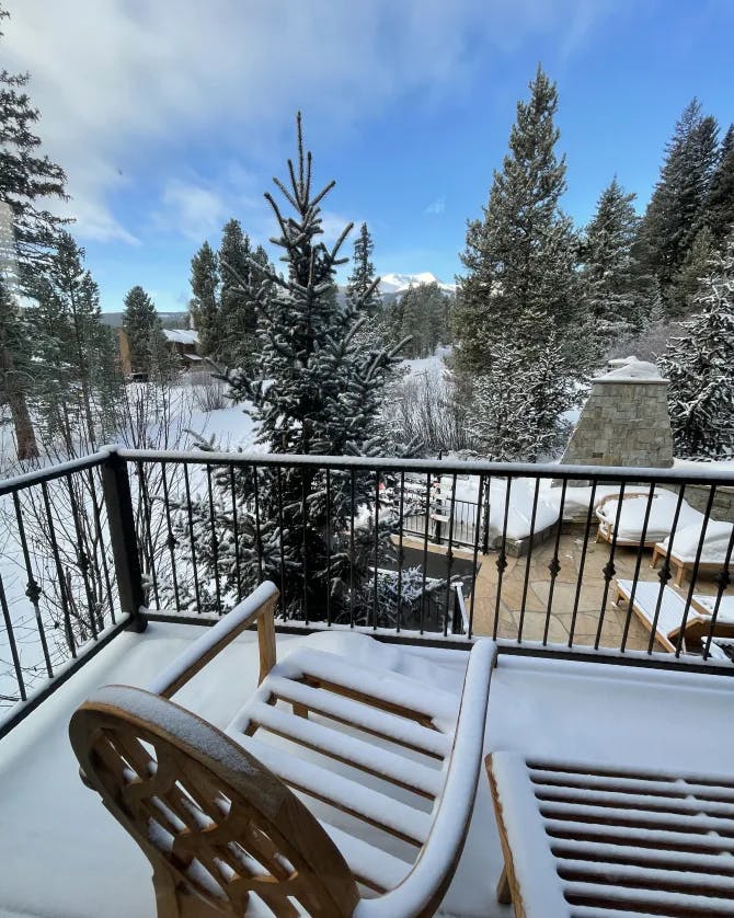 A snow covered chair and table on a balcony overlooking a snowy scenery with pine trees. 