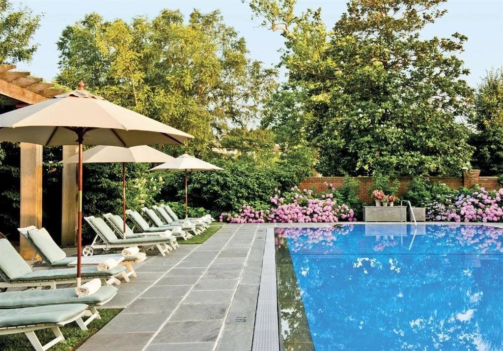 light-green loungers, green trees and hedges, and pink flowers surround a pool on a sunny day
