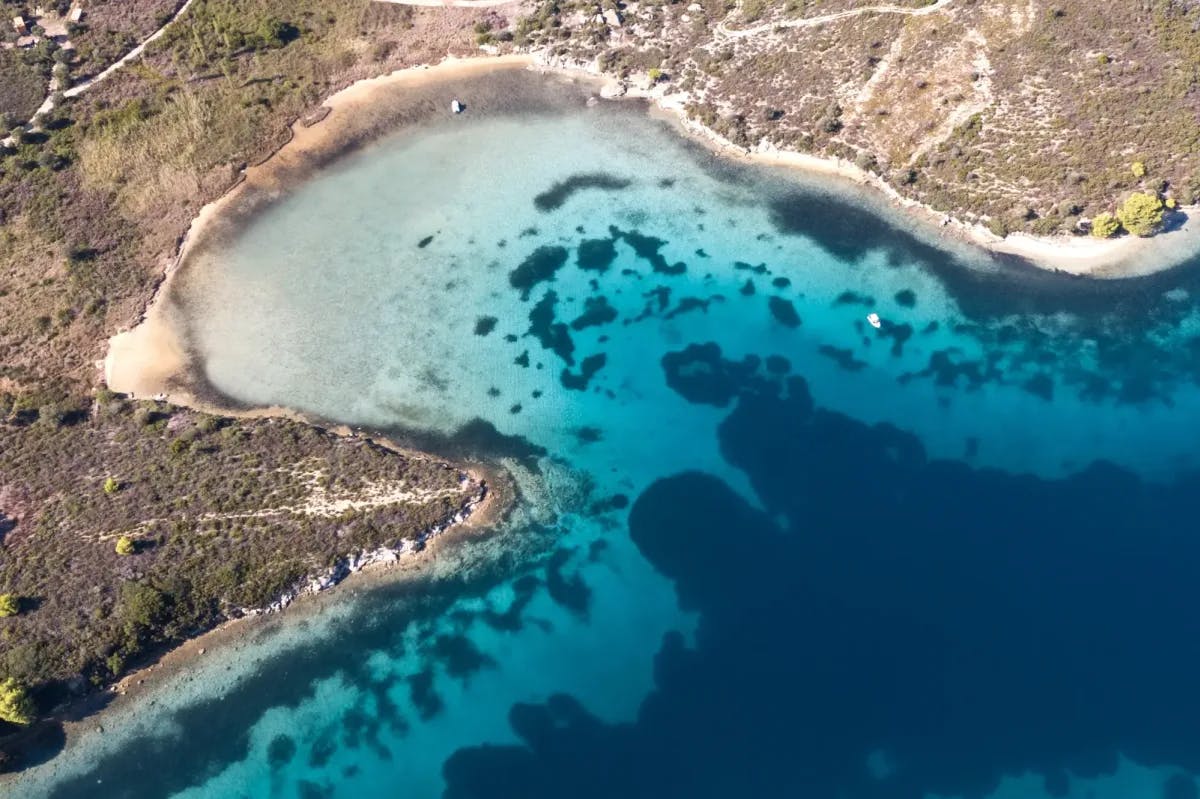Aerial view of a beautiful beach in Halkidiki, Greece. Rocky hills with sparse vegetation lead up to a volcanic beach and a mix of turquoise and sapphire waters