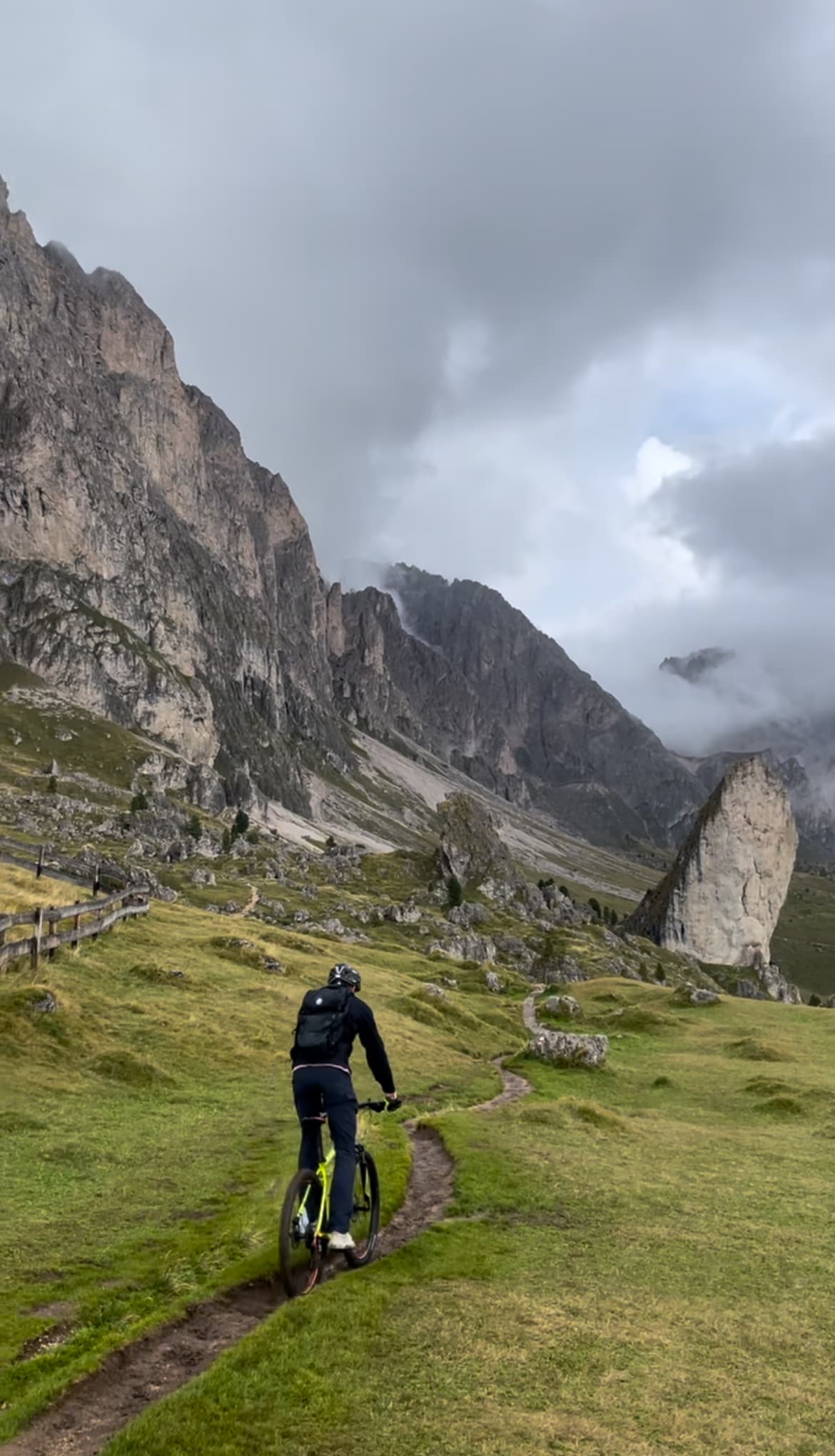 A person cycling on a grassy dirt path surrounded by mountains on a cloudy day
