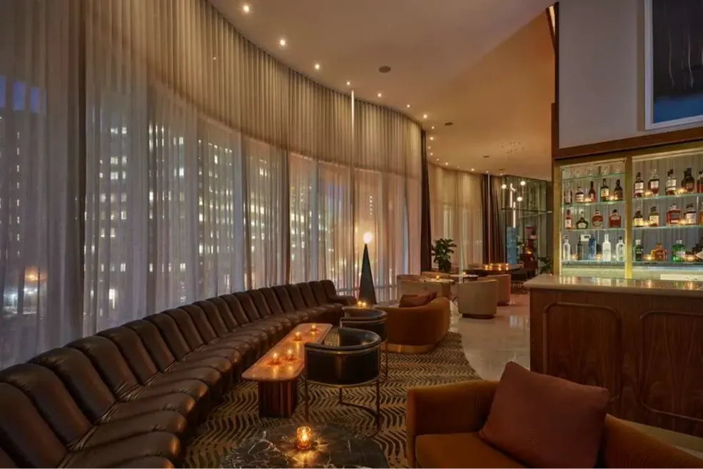 A classy, upscale lounge with retro vibes and huge floor-to-ceiling windows covered with thin drapes