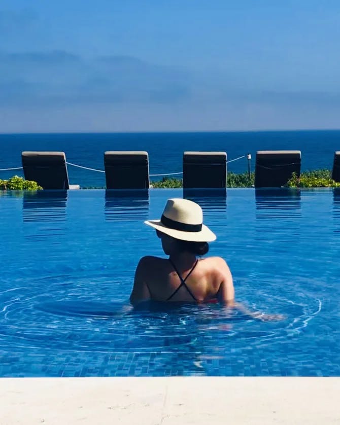Samantha wearing a hat and relaxing in a blue pool that overlooks the ocean