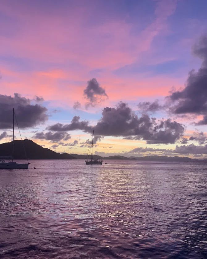 A beautiful purple and pink sunset over water with sailboats and a mountain in the distance