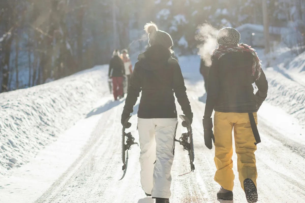 Two people wearing ski gear and walking down a snowy path with more people up ahead and pine trees in the distance. 