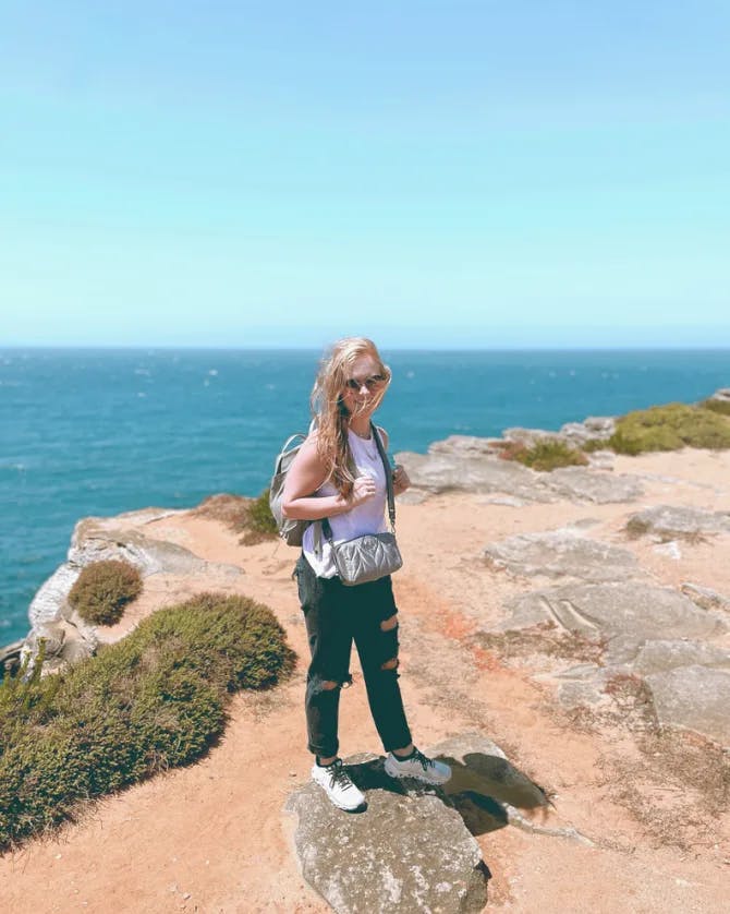 Picture of Merritt wearing a white top and blue jeans while standing on a rocky cliff with a view of the sea in the background on a sunny day 