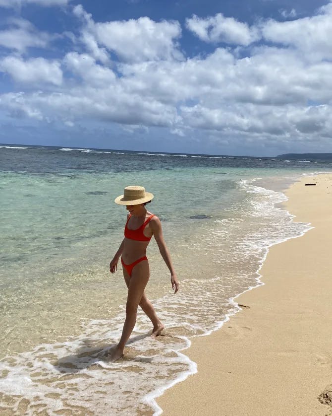 Gabby wearing a swimsuit and hat walking on the beach with the ocean and cloudy blue sky in the background