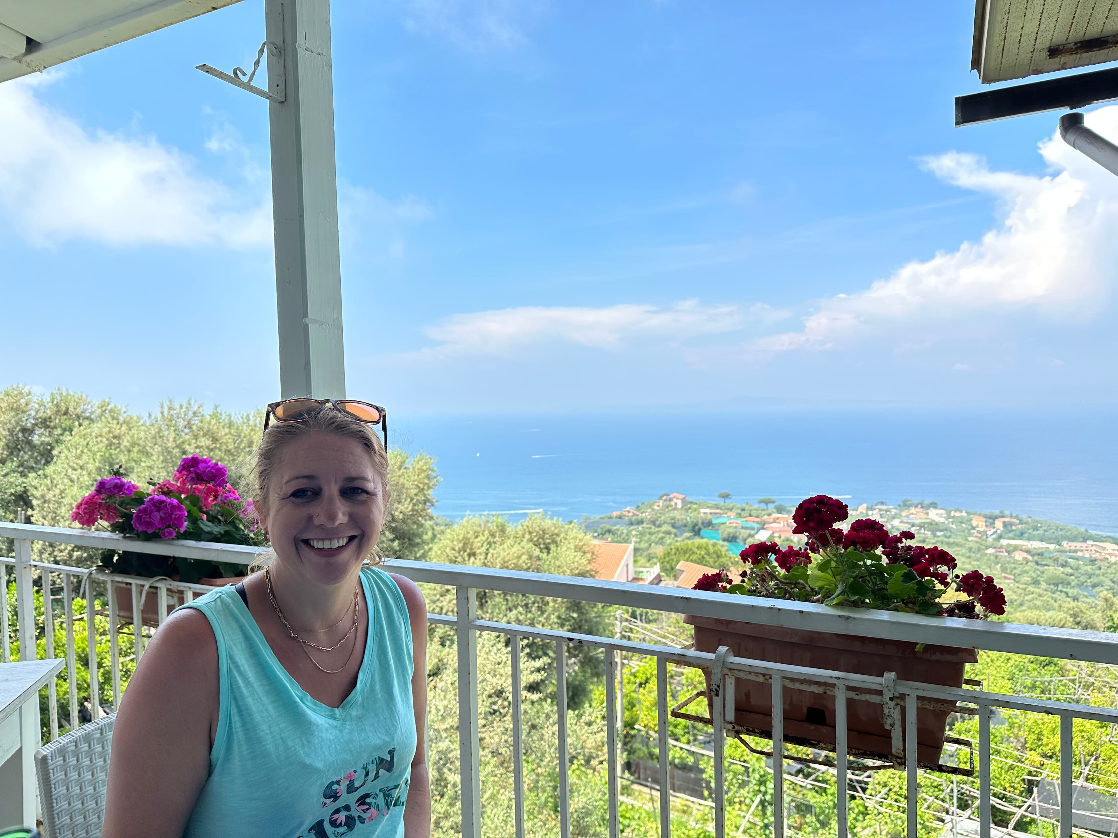 Jennie sitting on a balcony and smiling with a semi-tropical landscape and sea view in the background.