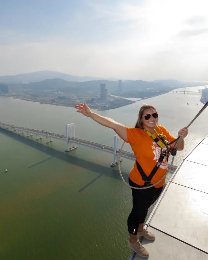 A picture of Ashley wearing an orange top and black pants preparing to bungee jump at Macau Tower Convention and Entertainment Center