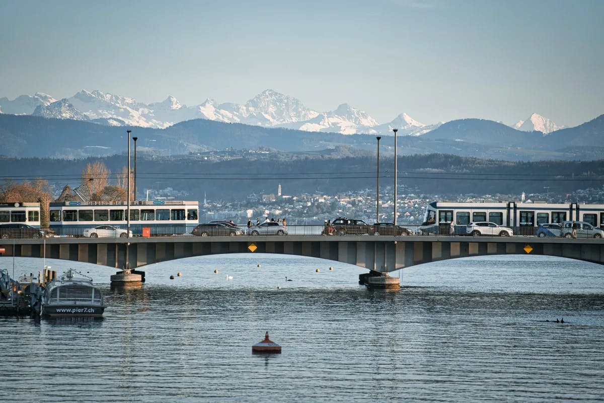 A picture of the bridge over a lake with boats during the daytime and a snow capped mountain range in the background. 